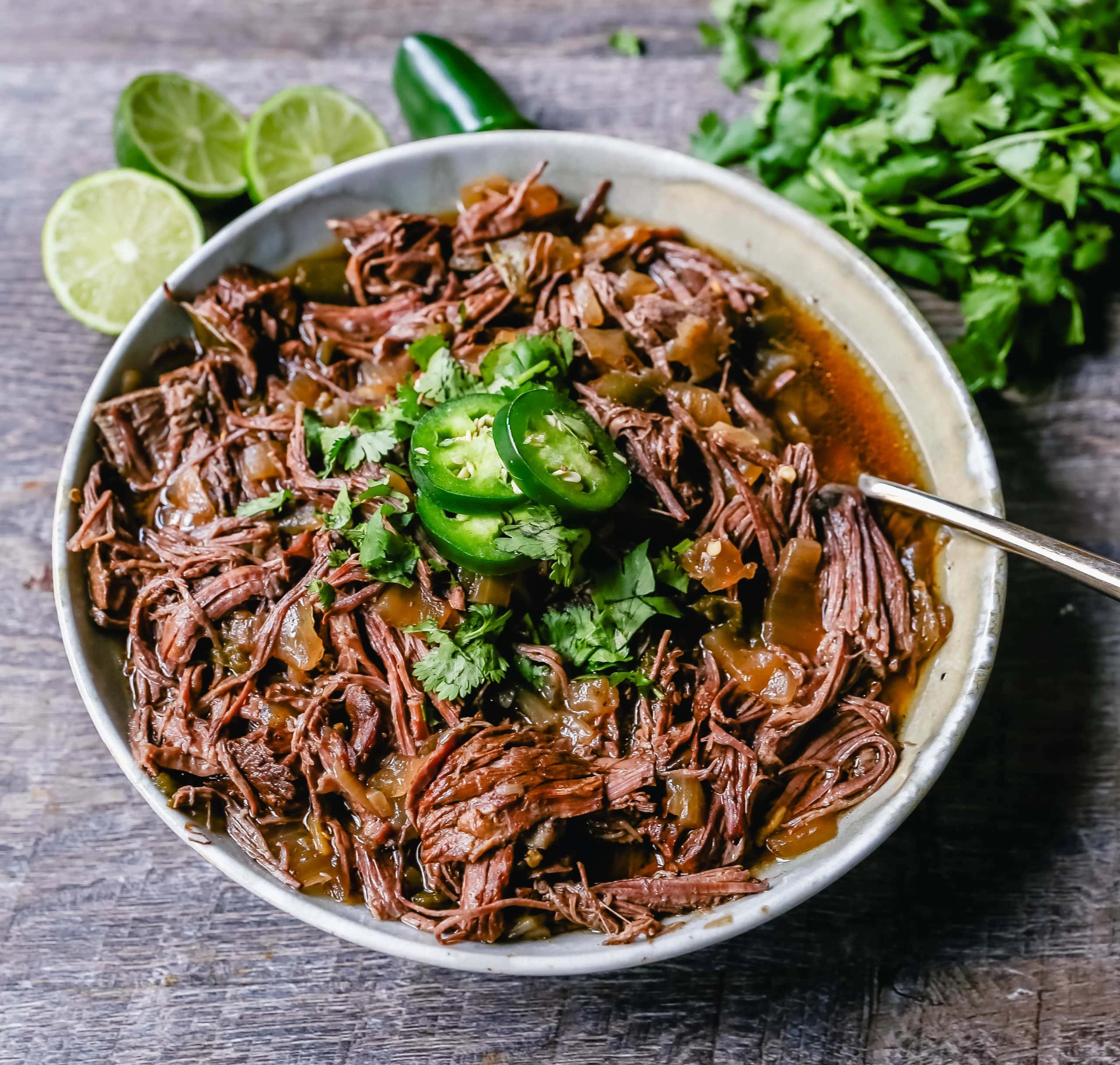 Bowl of shredded beef barbacoa topped with cilantro and jalapeño slices with lime wedges on the side