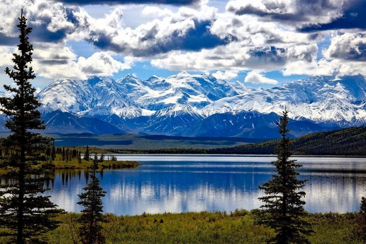 Snow-covered mountain range above a calm blue lake with evergreen trees in the foreground under dramatic clouds