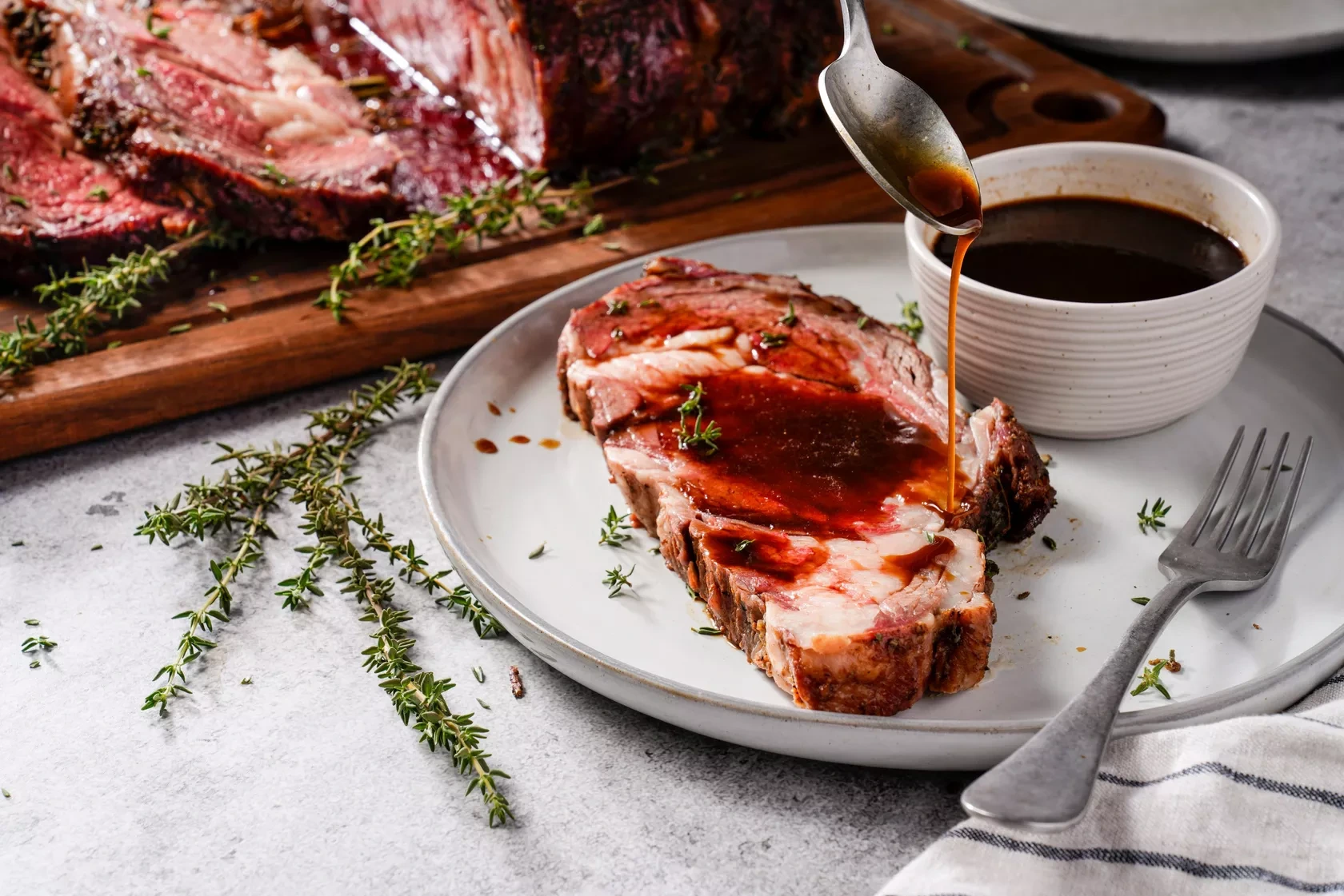 Sliced roast beef on a plate with au jus sauce being spooned over the meat and a bowl of dipping sauce on the side