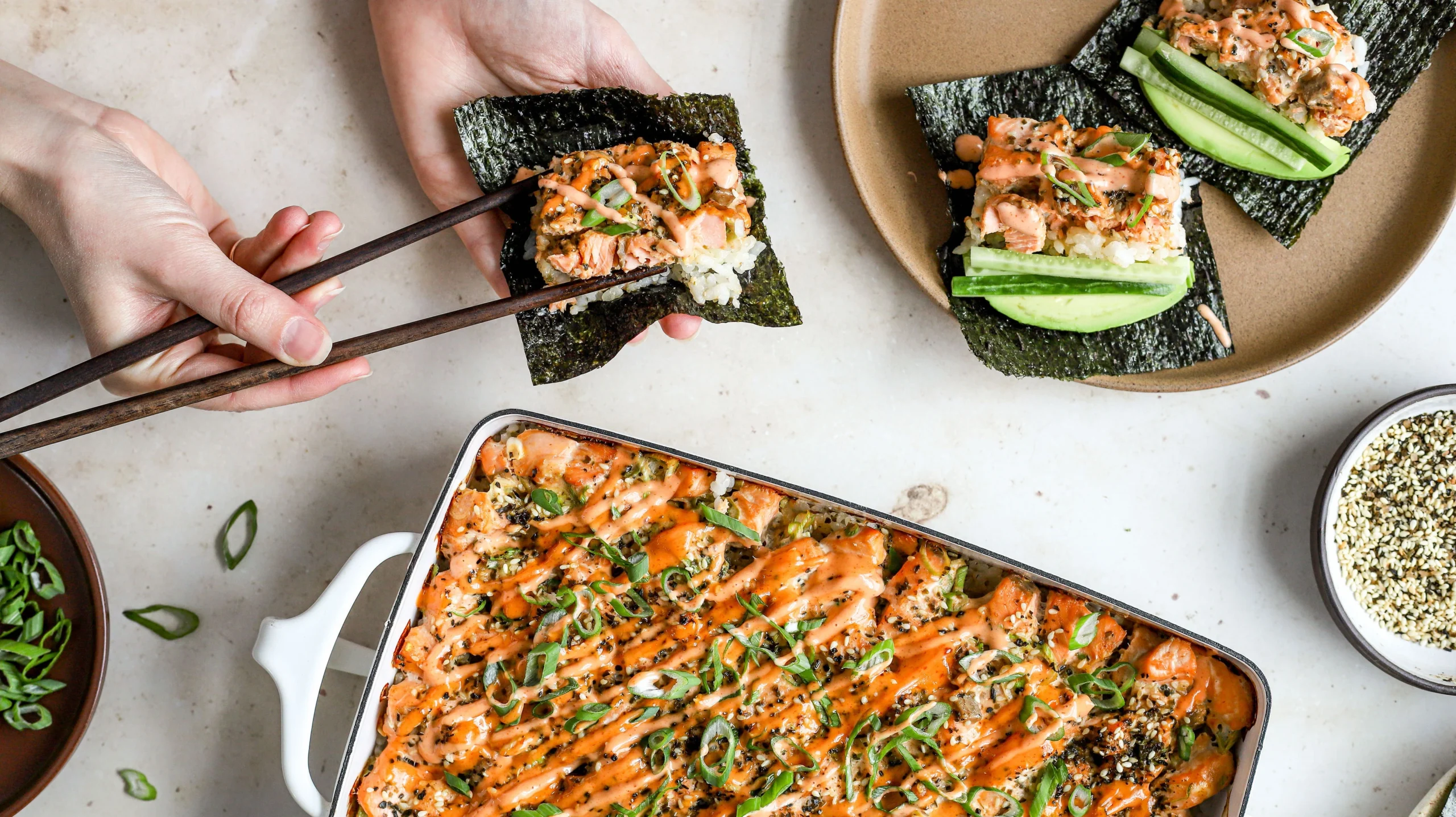 Sushi bake in a casserole dish topped with spicy mayo and green onions, served with nori sheets and cucumber slices